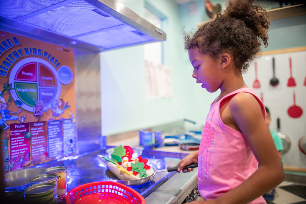 child wearing pink shirt and playing in kitchen play exhibit