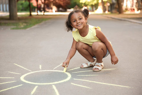 young child drawing sun with sidewalk chalk on asphalt