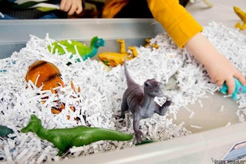 toddler playing with shredded paper in a bin full of dinosaur toys as a sensory activity