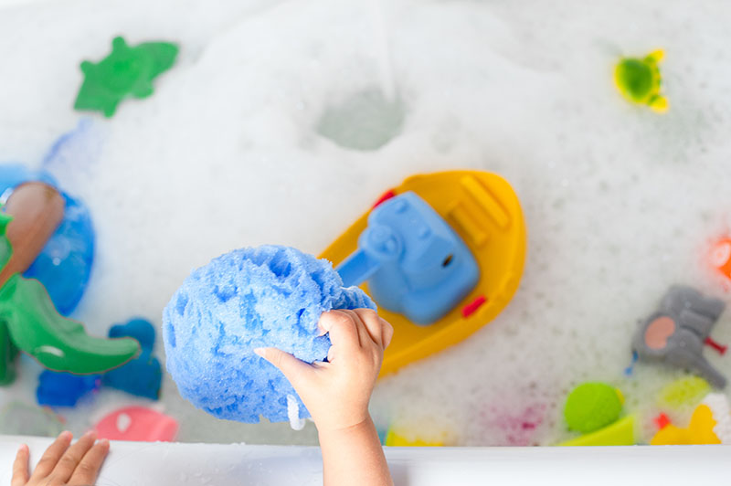 Manos de un niño sosteniendo una esponja azul con una bañera en el fondo