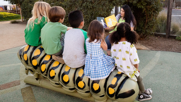 5 young children sitting on a large caterpillar bench at the Children's Museum of Sonoma County listening to a story being read aloud by an adult sitting in front of them