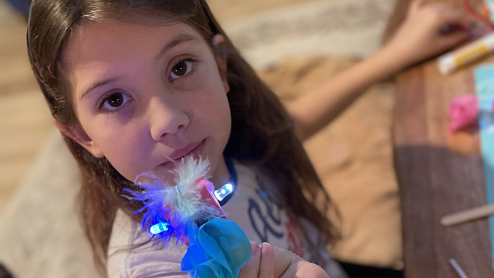 Young girl showing off her light-up accessory that she made after learning how to create an electric circuit with the Children's Museum's Electric Bling Kit and Caboodles activity box