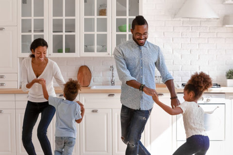 Happy parents and two toddlers dancing to music in kitchen, holding hands, having fun and laughing