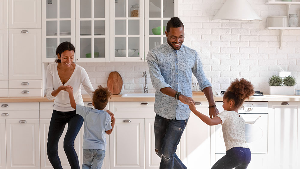 Happy parents and two toddlers dancing to music in kitchen, holding hands, having fun and laughing