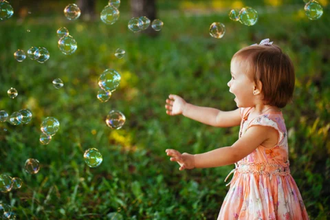 A little girl smiling and playing with soap bubbles outside with grass and trees in the background