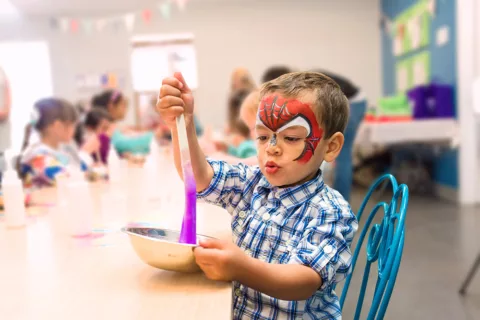 A toddler wearing Spiderman face paint making DIY slime during a Halloween-themed activity