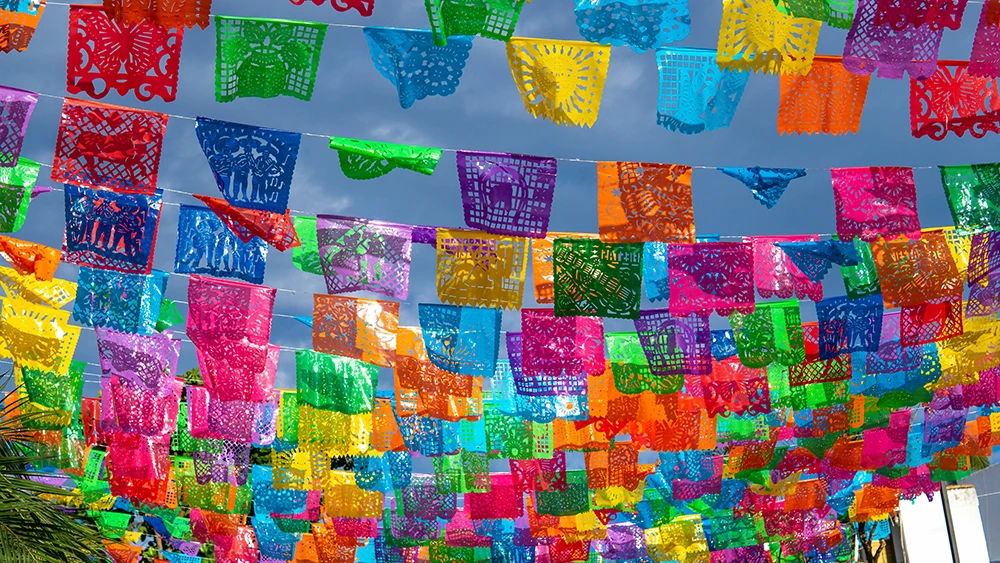 Several colorful Papel Picado banners hanging between two buildings with a sunny blue sky in the background.