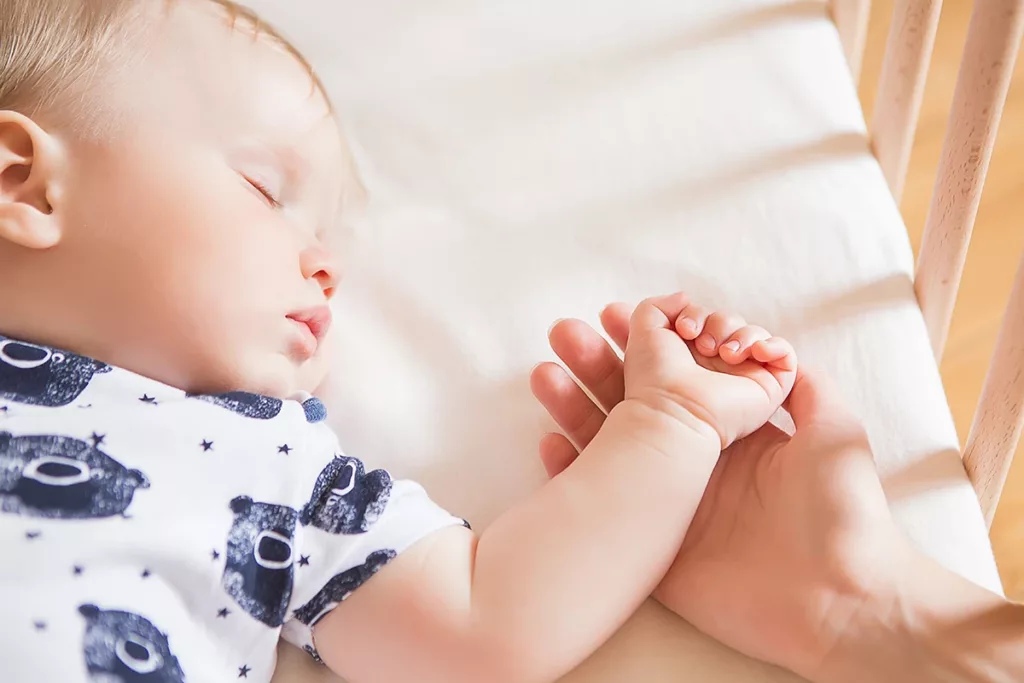 A baby, less than one years old, happily sleeping in their crib, while a parental figure holds their hand with love and care.