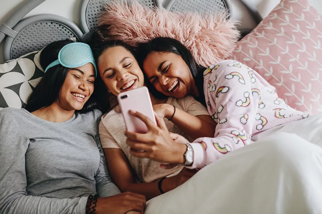 Teenage girls at a slumber party laughing and smiling while play on a smartphone together.