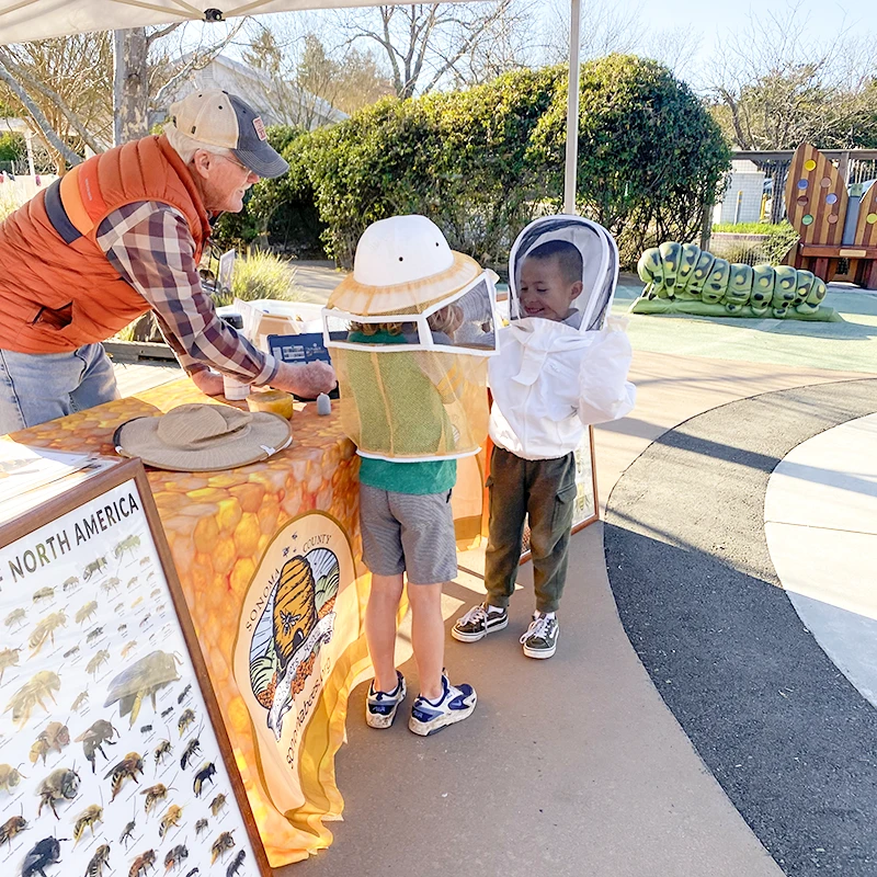Un apicultor de la Asociación de Apicultores del Condado de Sonoma y dos niños con trajes protectores para divertirse en el Museo de los Niños del Condado de Sonoma durante el programa educativo mensual para niños Meet a Beekeeper.