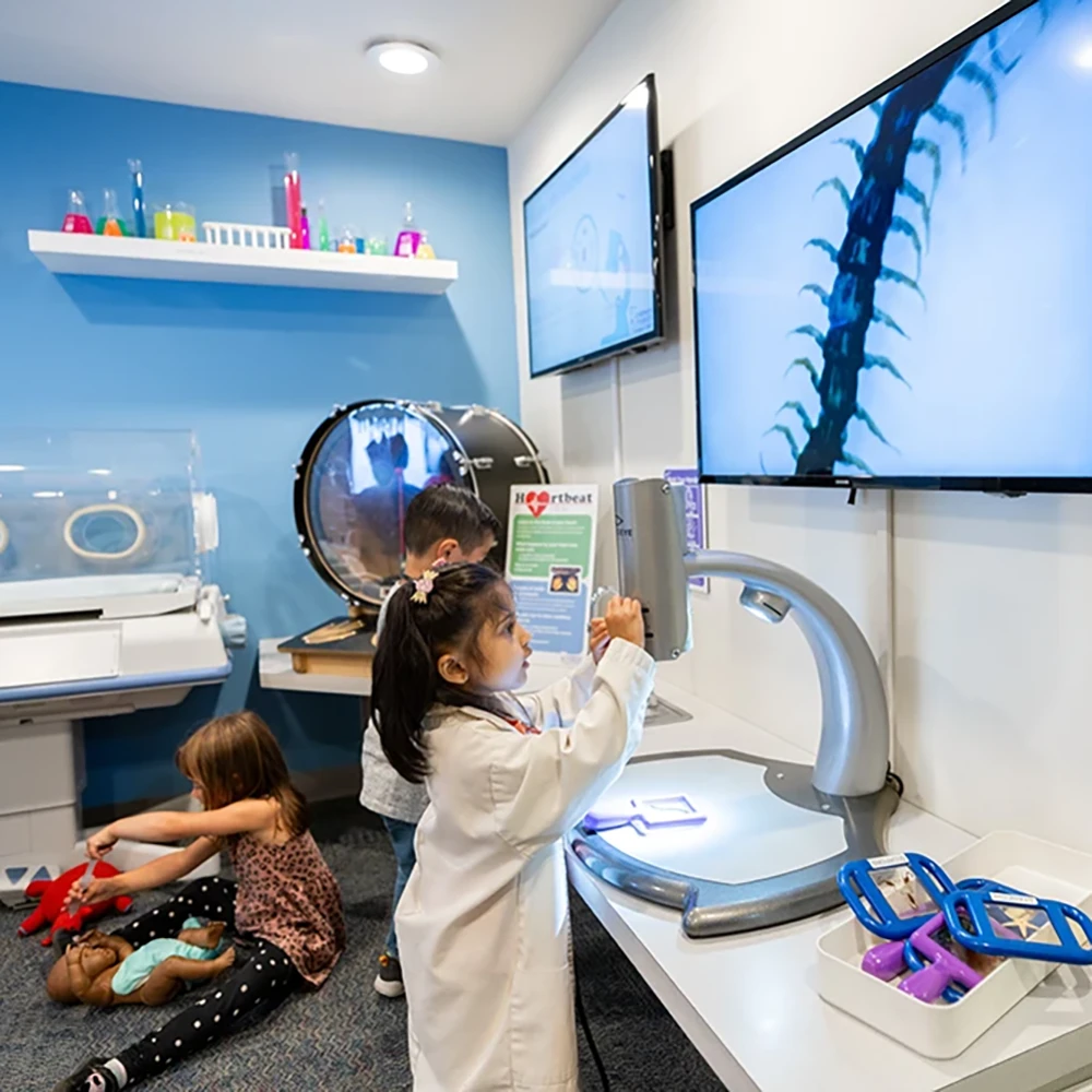 A child wearing a child-sized white lab coat laughing and playing with an interactive medical and science-themed exhibit called the Science & Medical Lab exhibit at the Children's Museum of Sonoma County in Santa Rosa, Ca.