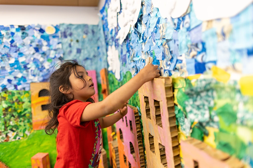 A child doing a large scale, collaborative art project in Ella's Art Studio featuring colorful pieces of paper covering 2 entire walls in the studio to create a beautiful scene with grass, the sky, the sun, flowers, and buildings.