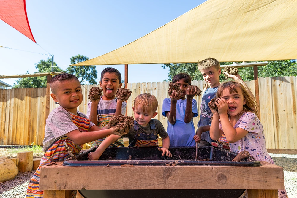 Un grupo de seis niños ríen y juegan juntos en una bandeja de barro sensorial, todos miran a la cámara y ponen caras divertidas mientras sostienen puñados de barro en la exposición Mud Lab del Children's Museum of Sonoma County de Santa Rosa, Ca.