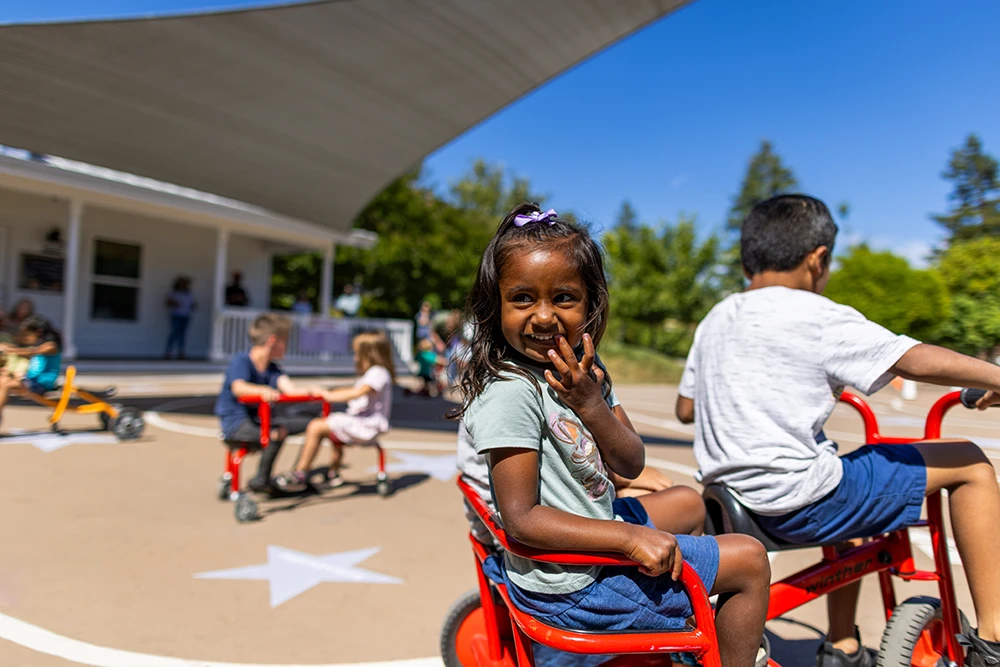 A young child sitting in the back seat of a 2-person children's tricycle looking backwards towards the camera and smiling excitedly while another child pedals the bike around the Bike Track.