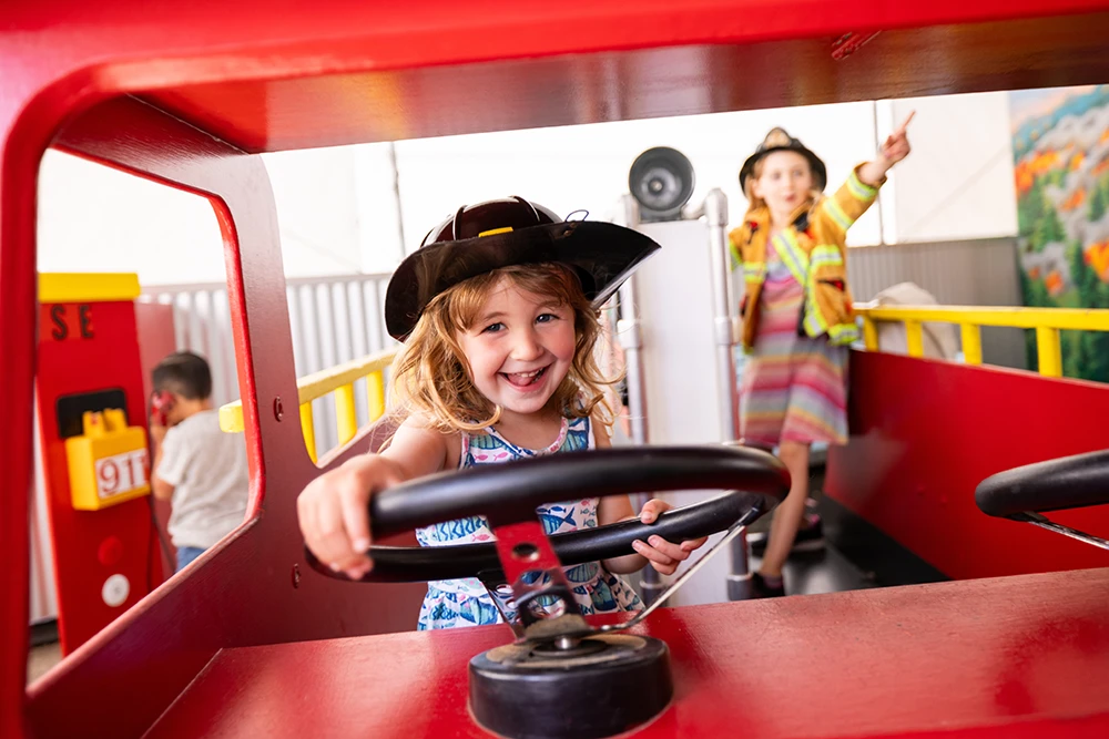 A child wearing a dress-up Fire Fighter helmet laughing and playing in a child-sized fire truck at the Children's Museum of Sonoma County in Santa Rosa, Ca.