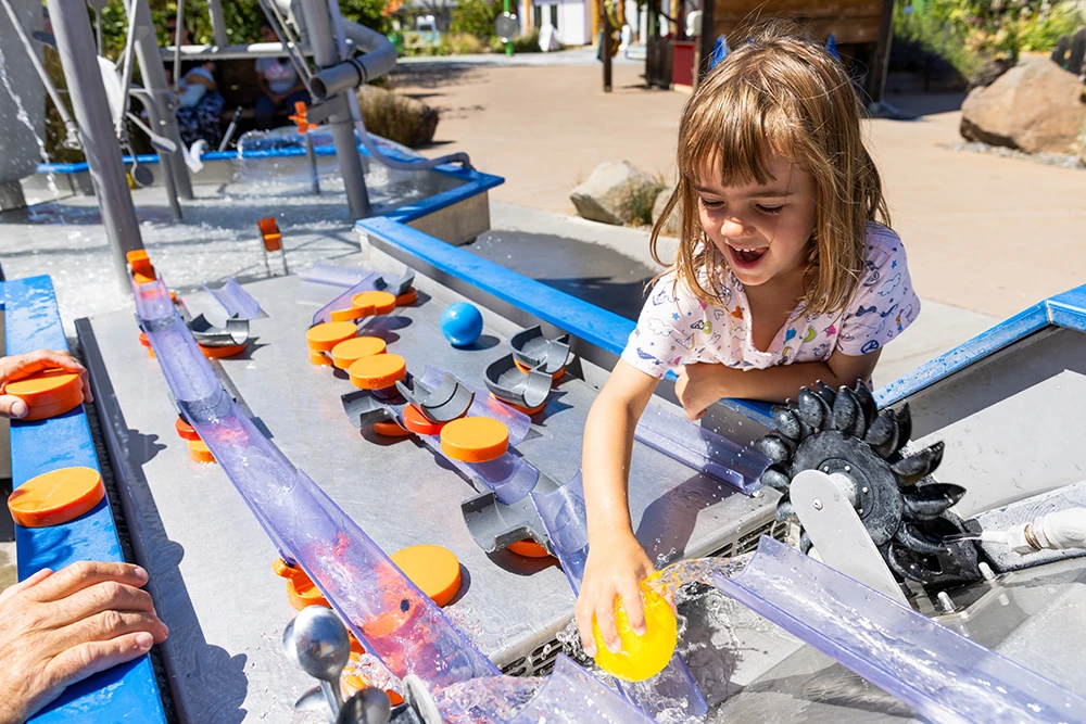 A child laughing and playing with plastic toy balls in an interactive water-themed exhibit called the Mechanical Waterway 2.0 exhibit.