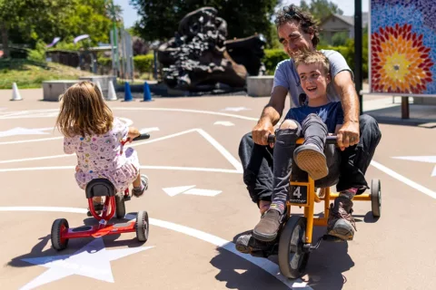 A child and their adult guardian laughing and playing together on the Bike Track at the Children's Museum of Sonoma County in Santa Rosa, Ca.