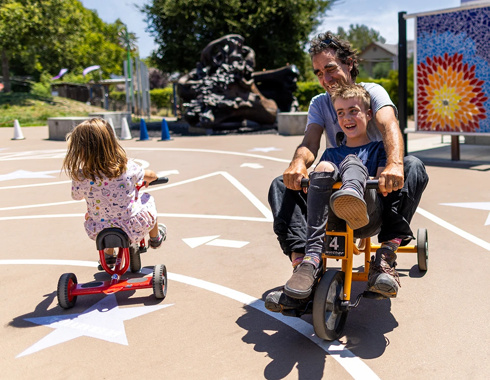 A child and their adult guardian laughing and playing together on the Bike Track at the Children's Museum of Sonoma County in Santa Rosa, Ca.