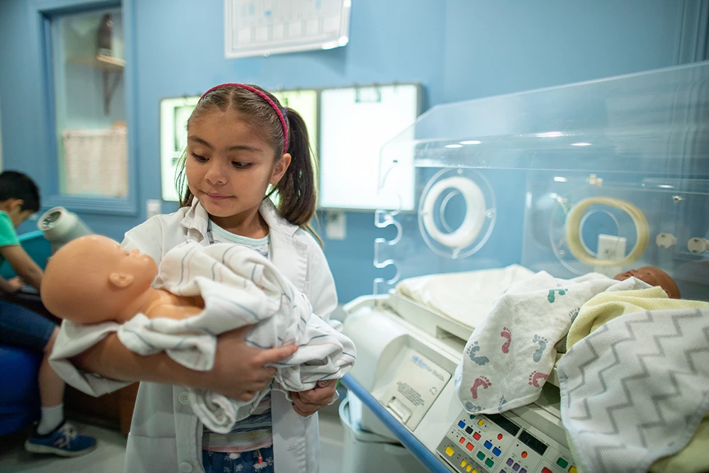 Un niño con una bata blanca de laboratorio de tamaño infantil sonríe mientras sostiene un muñeco bebé de juguete en una exposición interactiva de temática médica y científica llamada Science & Medical Lab.
