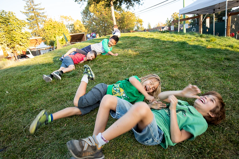 Un grupo de cuatro niños ríen y ruedan por una colina cubierta de hierba en el Jardín de Mary del Museo Infantil del Condado de Sonoma en Santa Rosa, Ca.