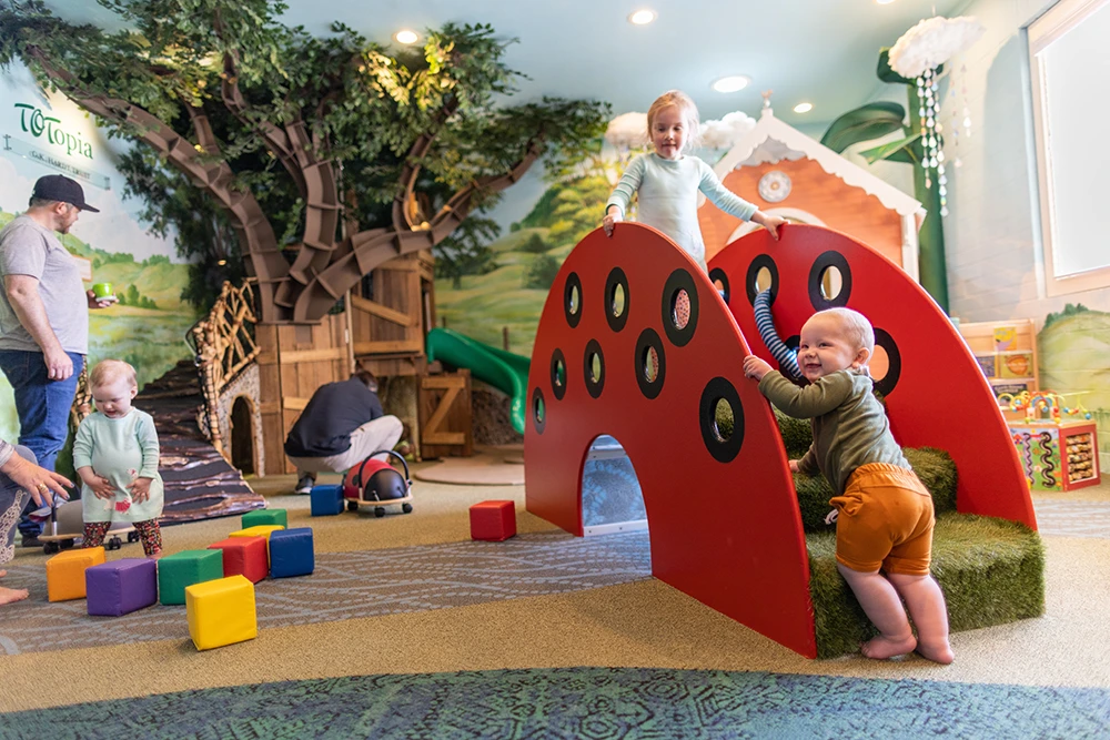 Three toddlers and their adult guardians laughing and playing in Totopia. An area in the Children's Museum specifically dedicated to children under 2 years old.