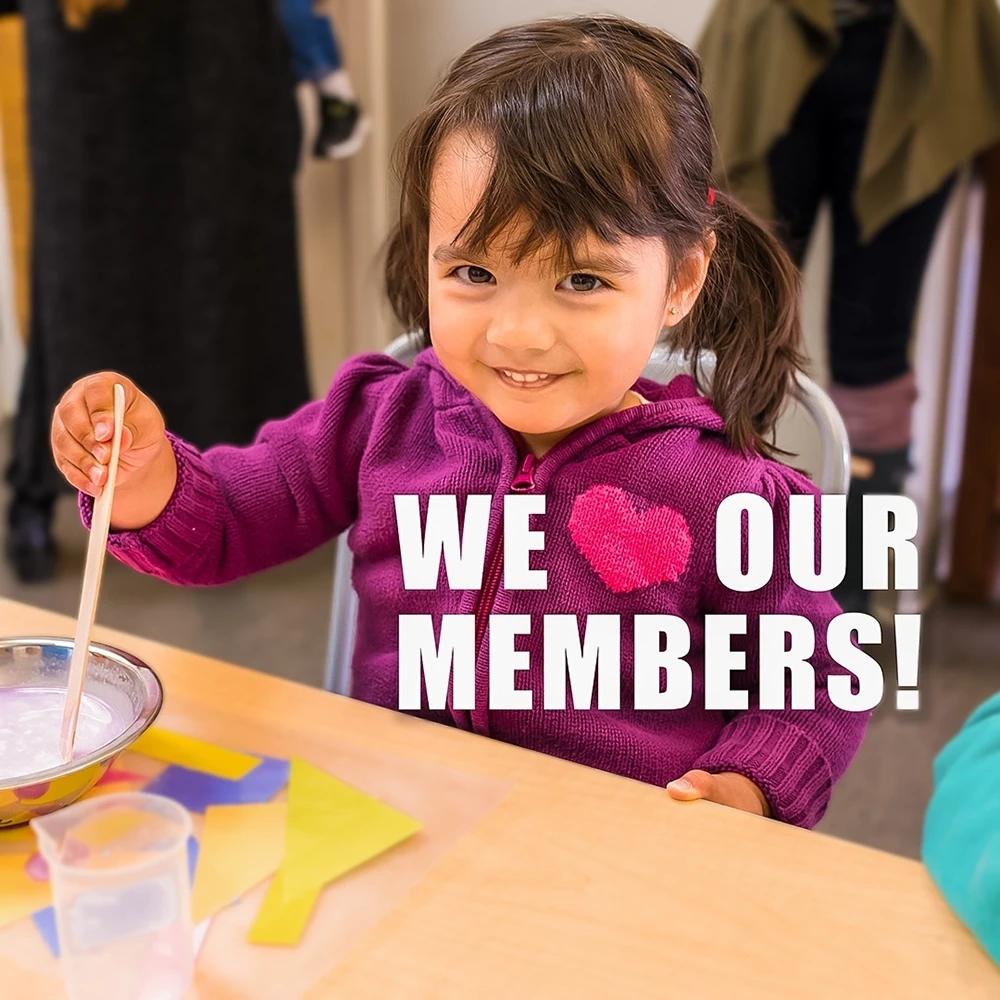 A child sitting at a table doing an arts and crafts project in Ella's Art Studio at the Children's Museum of Sonoma County in Santa Rosa, Ca. She is smiling and looking at the camera. Her purple jacket has a pink heart design on it. There is text overlay the picture that reads: "We Heart our Members!" The heart on her jacket is being used to represent the "Heart" in the overlay text.