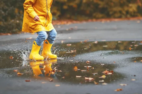 A young child, toddler, wearing a yellow raincoat and yellow rain boots, happily jumping in a puddle, splashing rain water over the sidewalk, and enjoying a nice walk as an outdoor rainy day activity.