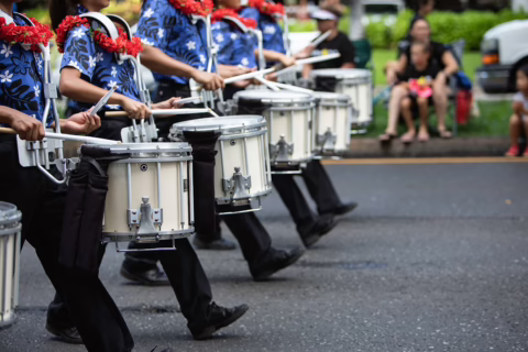 A line of drummers in a Marching Band, walking down a road participating in a Sonoma County Parade as people, families, and children, happily watch from the sidewalk