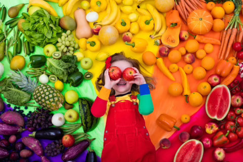 a young child surrounded by colorful fruits, veggies, and rainbow colors, smiling and hold two apples up to her eyes so it looks like she has silly apple eyes; teaching children where food comes from concept