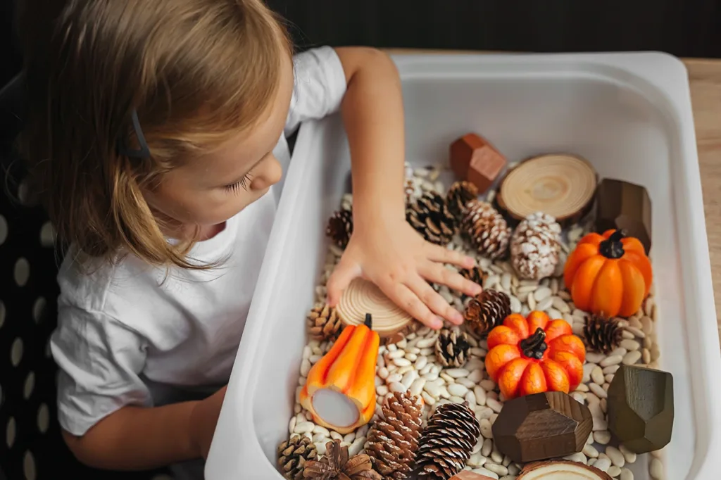 Vista lateral de un niño pequeño jugando con un cubo sensorial de otoño lleno de pequeñas calabazas de imitación, piñas y judías secas, que sirve como ejemplo visual de las ideas de juego sensorial de otoño para niños pequeños.