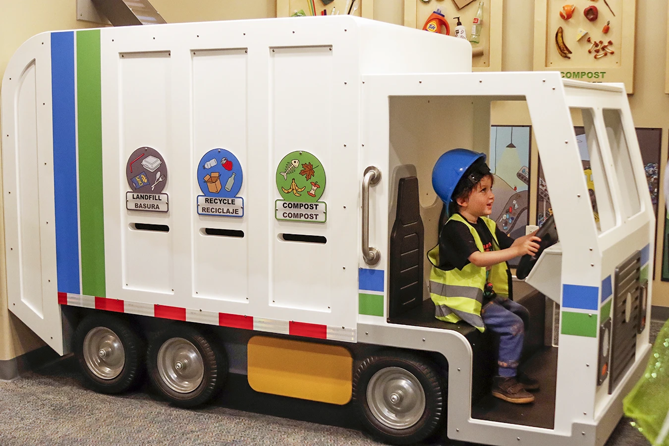 Un niño pequeño vestido con un casco y un chaleco de seguridad de juguete fingiendo conducir un camión de basura estilo Recology para niños, con las palabras "Landfill/Basura", "Recycling/Reciclaje" y "Compost/Compost" escritas en los laterales del camión, en la exposición Sorting Station del Museo Infantil del Condado de Sonoma.