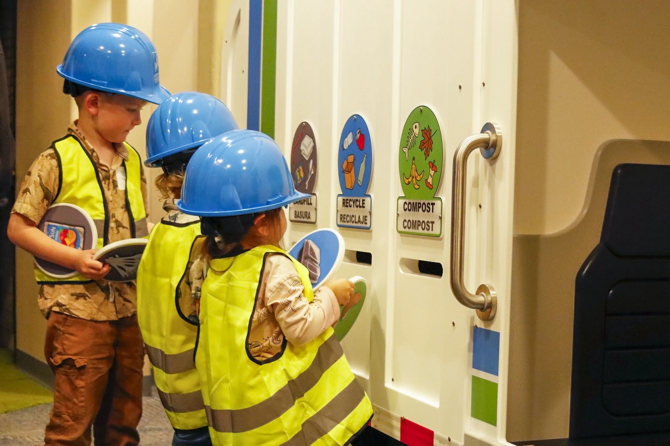 Tres niños pequeños vestidos con cascos y chalecos de seguridad de juguete clasifican materiales de juguete en compartimentos etiquetados como "Basura", "Reciclaje" y "Compost" en la exposición «Estación de clasificación» del Museo Infantil del Condado de Sonoma.