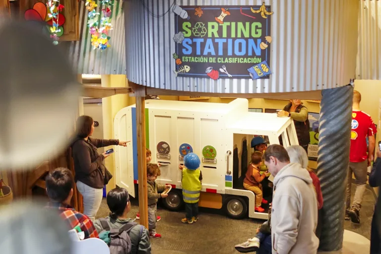 Niños jugando en la exposición "Sorting Station" (Estación de clasificación) del Museo Infantil del Condado de Sonoma, bajo la supervisión de adultos. Un letrero sobre la entrada de la exposición reza: «Sorting Station, Estación de Clasificación»."