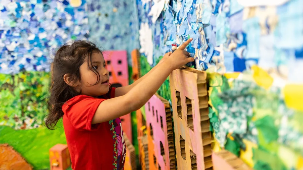 Un niño pequeño juega en el Museo Infantil del Condado de Sonoma, en una exposición cubierta de coloridos collages de papel que representan el cielo azul, las colinas verdes y la hierba, y los edificios de la ciudad hechos con cartón reciclado.