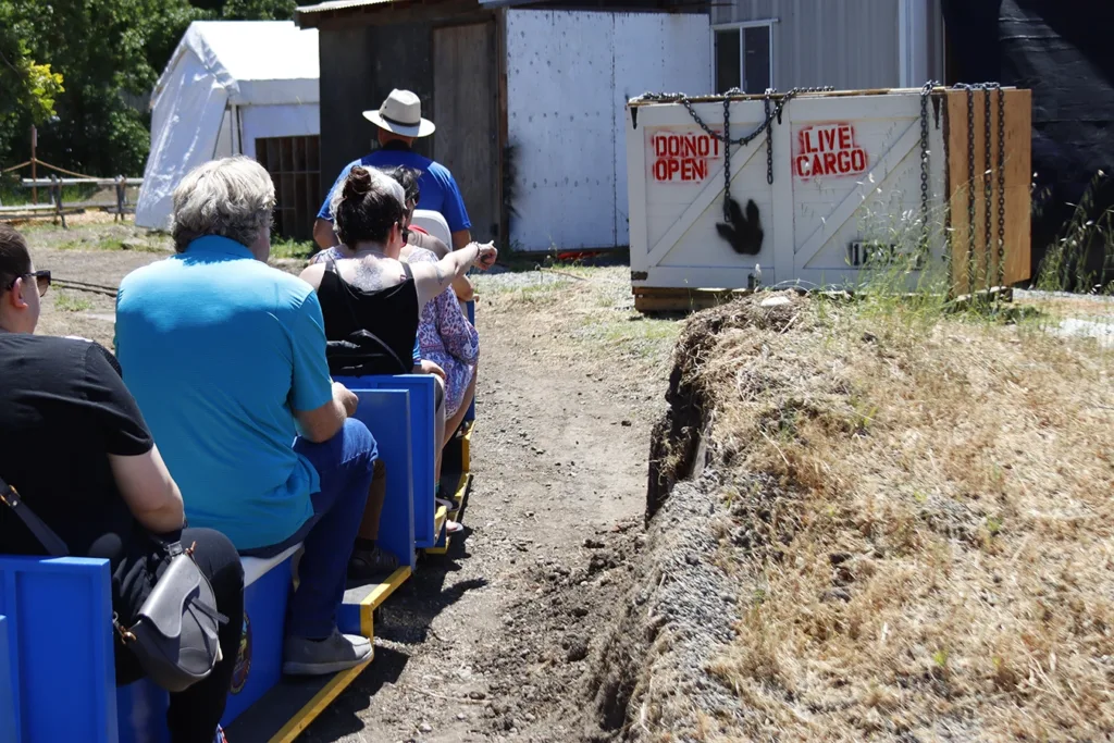 Children and caregivers riding a miniature train ride conducted by a Redwood Empire Live Steamers club member with one rider pointing to a giant shipping box to the side of the train trax that reads "DO NOT OPEN" and "LIVE CARGO" indicated there is a live dinosaur inside. Picture taken at the 2025 Dino Trax event at the Children’s Museum of Sonoma County in Santa Rosa, CA.