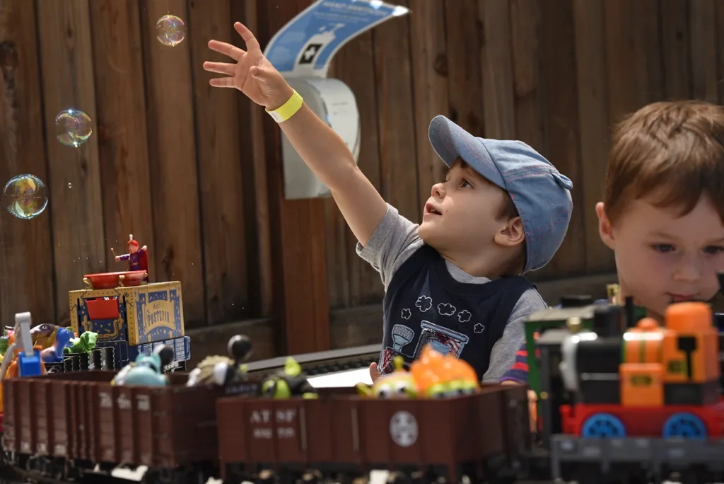 2 young children playing at the Children's Museum of Sonoma County's annual train and dinosaur themed event called, "Dino Trax!" One child is playfully reaching for a bubble that is floating by, while the other plays intently with a model toy train.
