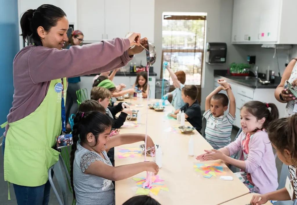 Un grupo de niños y miembros del personal del museo sentados alrededor de una mesa grande, preparando slime colorido de manera artesanal en el Estudio de Arte de Ella, en el Museo Infantil del Condado de Sonoma.