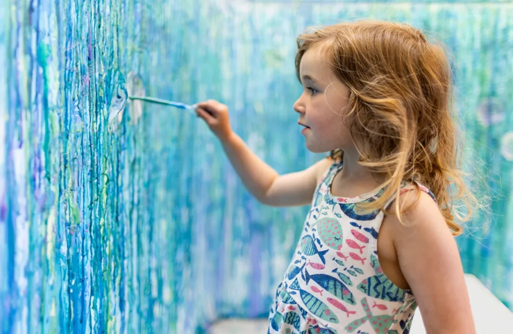 Un niño pequeño utiliza un pincel para pintar en una pared como parte de un proyecto artístico colaborativo en el Estudio de Arte de Ella, en el Museo de los Niños del Condado de Sonoma.