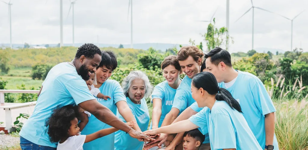 Varias familias, sonrientes y con camisetas de voluntarios, están de pie muy juntas formando un círculo, con las manos superpuestas, haciendo un grito de ánimo que simboliza el voluntariado familiar y las obras de caridad.