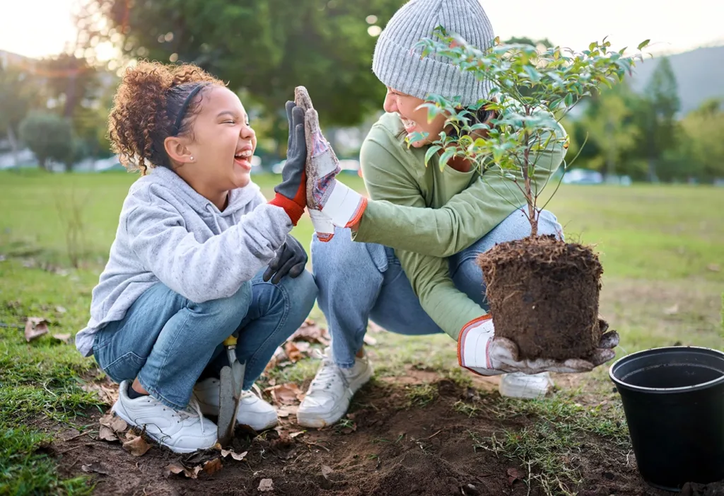 Un niño pequeño y su cuidador se dan un choque de manos mientras plantan un árbol en un parque local, como símbolo del voluntariado familiar y las obras de caridad.