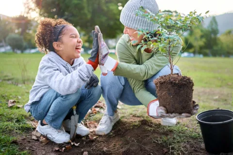 A young child and caregiver high-five as they plant a tree at a local park symbolizing family volunteering and charitable acts.