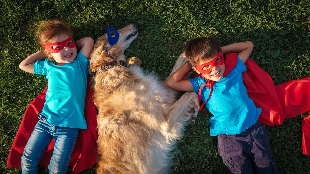 Two young children and their dog dressed up as superheroes laying in the grass.