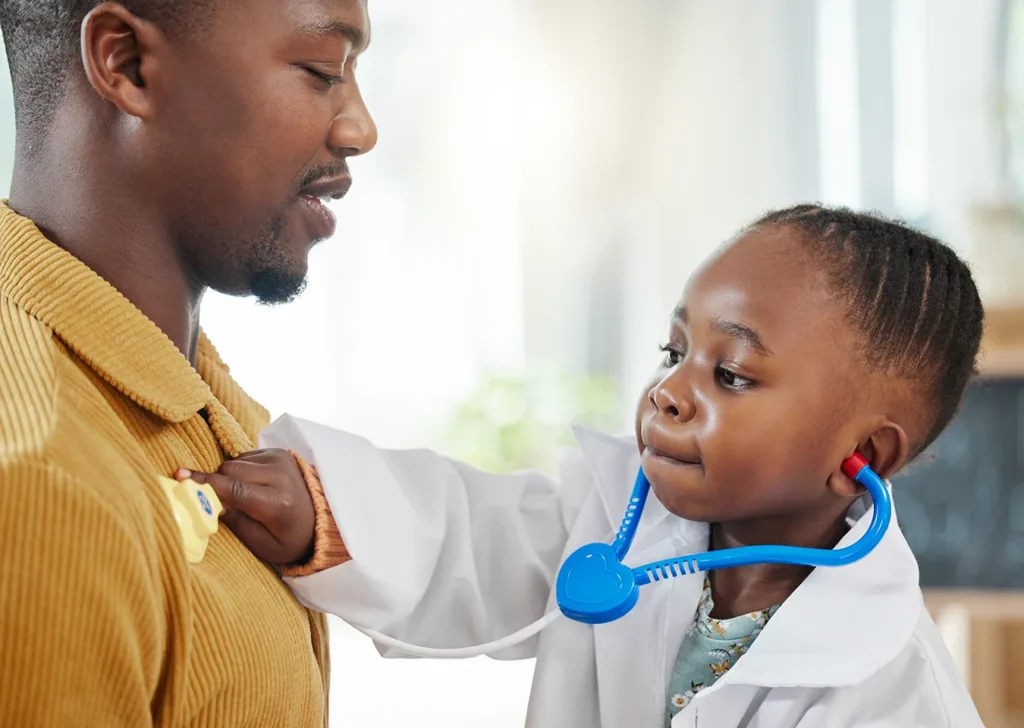 A young child dressed as a doctor using a toy stethoscope to pretend to listen to their father’s heartbeat.